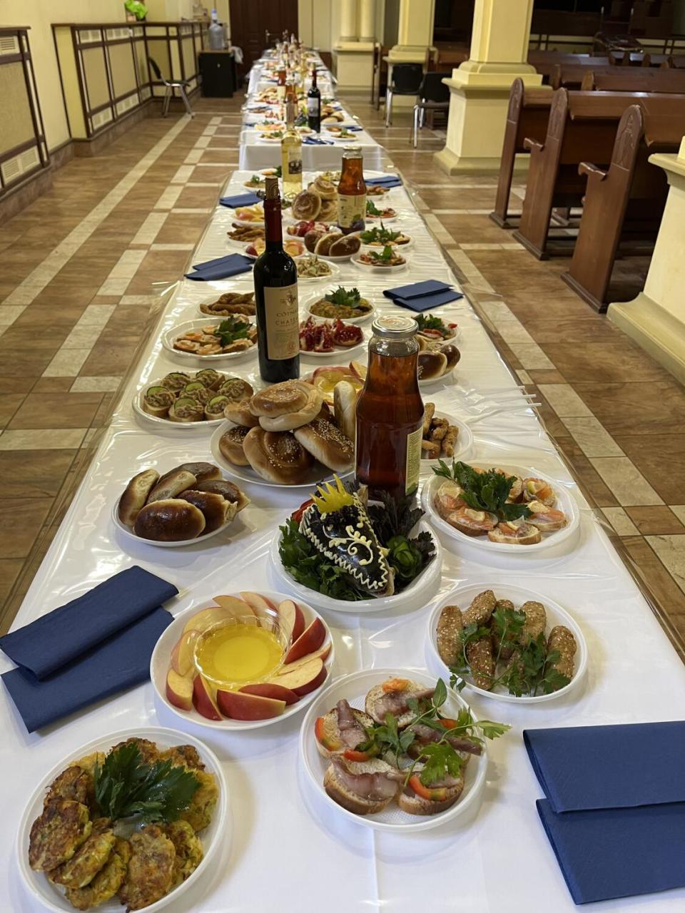 A long table, with white tablecloth and blue napkins, laden with plates of food and bottles of wine
