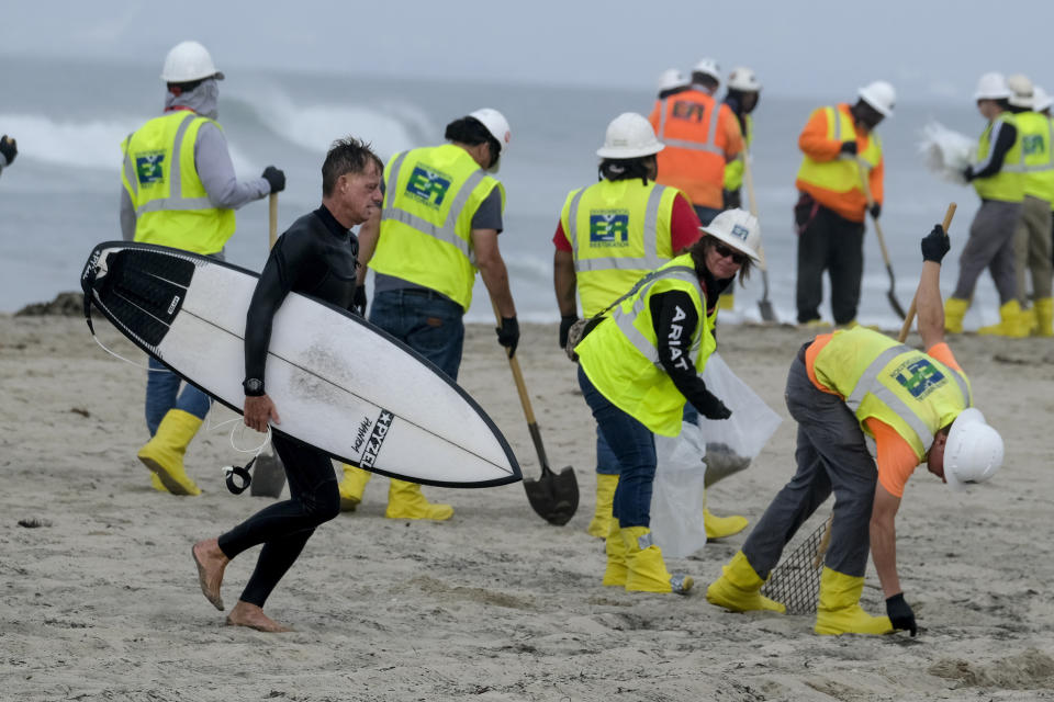 FILE - A surfer walks by as workers in protective suits continue to clean the contaminated beach in Huntington Beach, Calif., on Oct. 11, 2021. A pipeline operator and two subsidiaries have agreed to plead guilty to negligently discharging oil off the Southern California coast in connection with a pipeline break that covered beaches with blobs of crude. (AP Photo/Ringo H.W. Chiu, File)