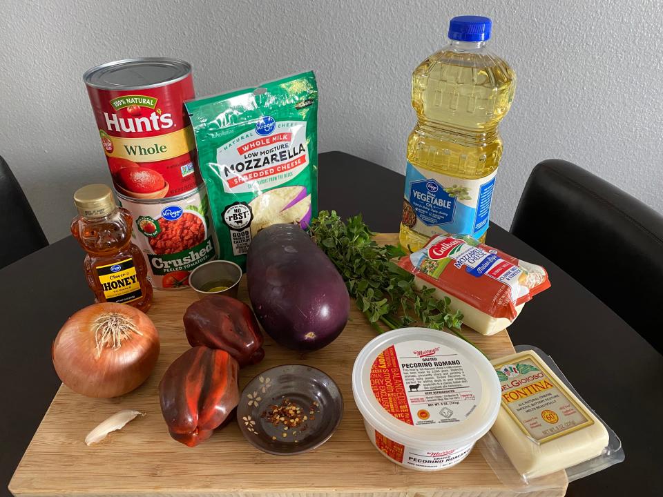 An assortment of ingredients on a cutting board, including an onion, an eggplant, honey, mozzarella cheese, olive oil, and red pepper flakes.