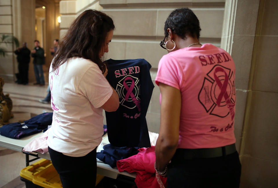 SAN FRANCISCO, CA - MARCH 26:  Former San Francisco firefighter and cancer survivor Denise Elarms (R) looks at a S.F.F.D. t-shirt with a pink cancer ribbon during a remembrance ceremony held for San Francisco firefighters who have died of cancer on March 26, 2014 in San Francisco, California. Over two hundred pairs of boots were displayed on the steps inside San Francisco City Hall to symbolize the 230 San Francisco firefighters who have died of cancer over the past decade. According to a study published by the National Institute for Occupational Safety and Health, (NIOSH)  findings indicate a direct correlation between exposure to carcinogens like flame retardants and higher rate of cancer among firefighters.  The study showed elevated rates of respiratory, digestive and urinary systems cancer and also revealed that participants in the study had high risk of mesothelioma, a cancer associated with asbestos exposure.  (Photo by Justin Sullivan/Getty Images)