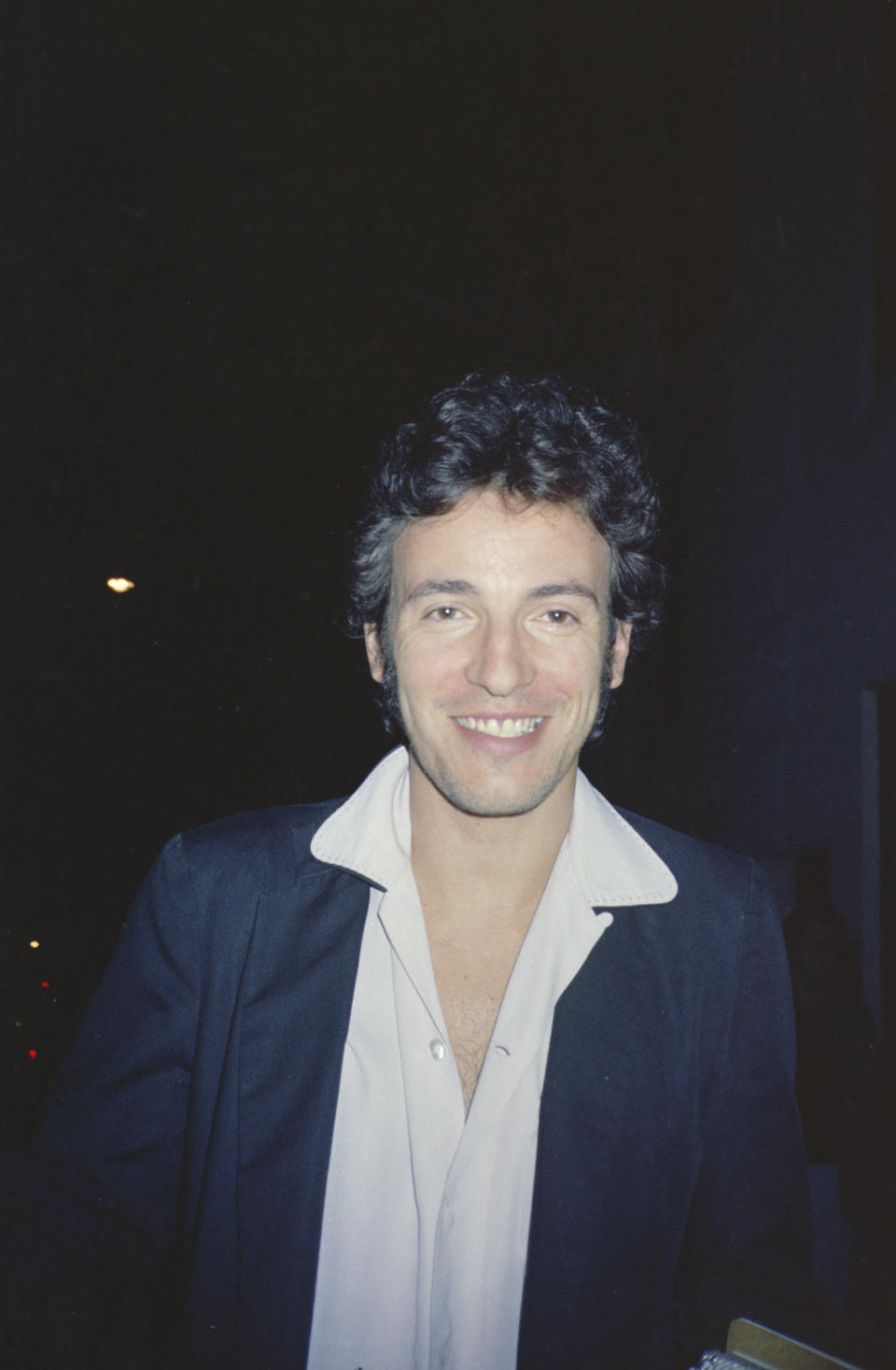 Bruce Springsteen smiles for a photo while wearing a white shirt and a dark jacket