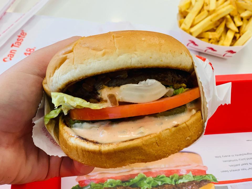 a hand holding in-n-out animal style burger in front of red tray and white table