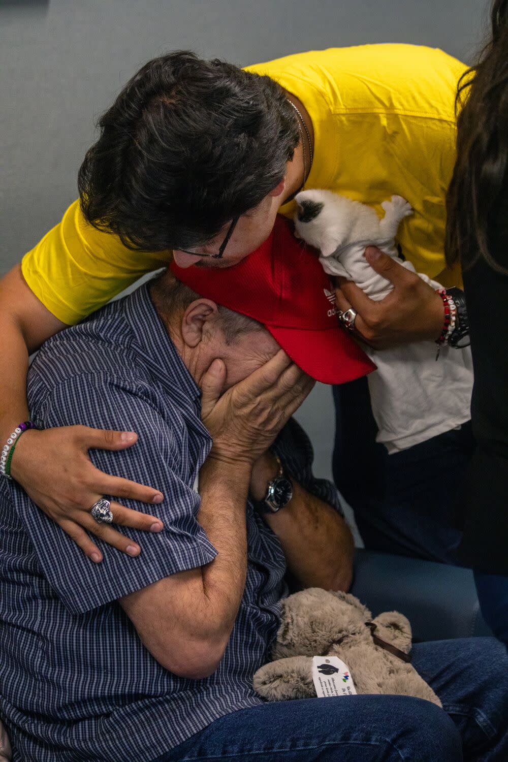 A young person bends down to embrace a seated man who is hiding his face in his palms.