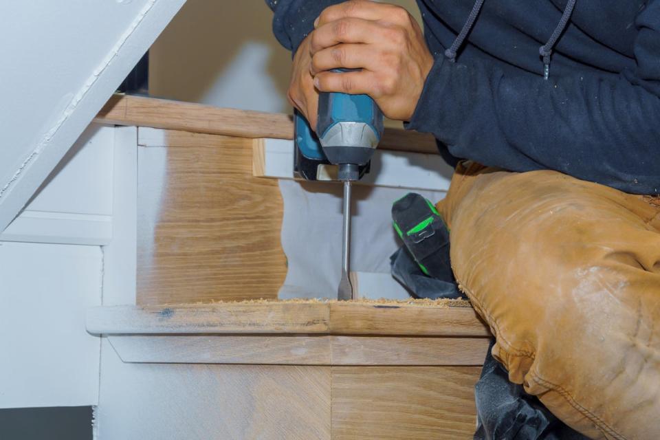 Man screws tread into riser with drill in wooden stairs.