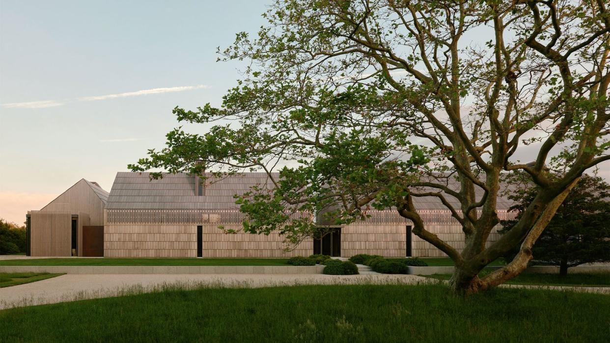 a long modern house with a side building made of cedar shingles and pitched roof, grass with a large tree, a gravel driveway and walkway with shrubs to house