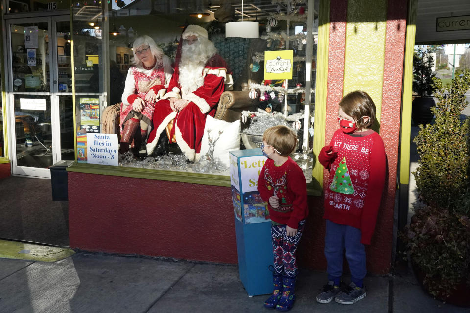 Beckett, right, and Hudson Gravley stand on the sidewalk in front of a storefront to pose for a photo with Santa and his wife, Mrs. Claus, in Bellingham, Wash., on Dec. 12, 2020. The storefront Santa sits on Saturday's during this holiday season in the shop window, where children can drop letters in a box outside and pose for photos as Santa sits inside behind. In this socially distant holiday season, Santa Claus is still coming to towns (and shopping malls) across America but with a few 2020 rules in effect. (AP Photo/Elaine Thompson)