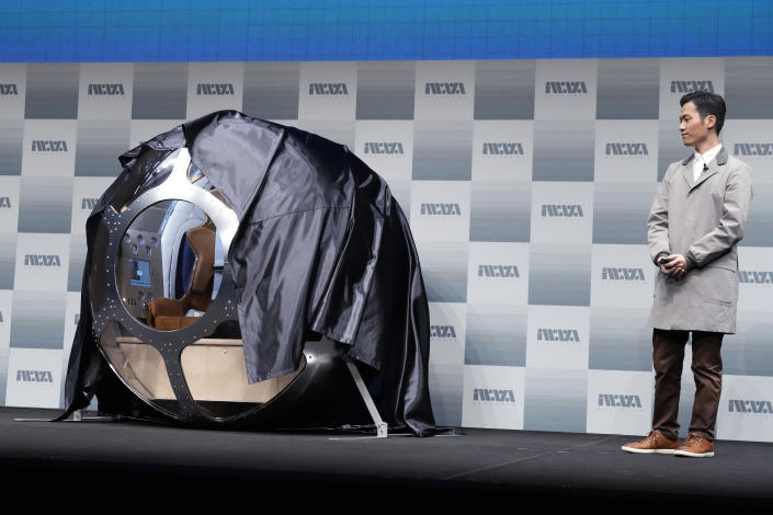 Keisuke Iwaya stands next to a steel orb with a beige floor and a chair facing an instrument panel, all partially covered by a shiny black plastic sheet.