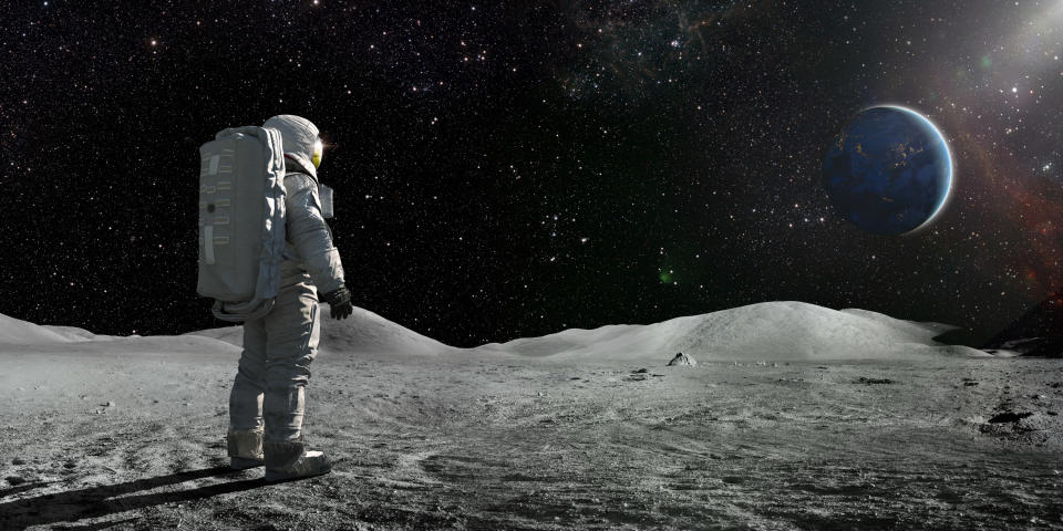 A lone astronaut standing facing away from the camera dressed in full space suit with backpack, stands still looking towards a distant planet Earth. The sun illuminates a side of Earth and hundreds of stars are visible in deep space.