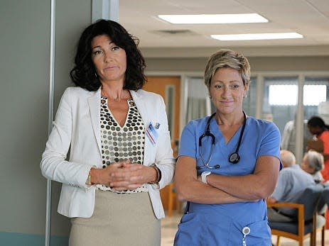 A woman with dark hair wearing a white blazer stands next to a blonde nurse in scrubs.