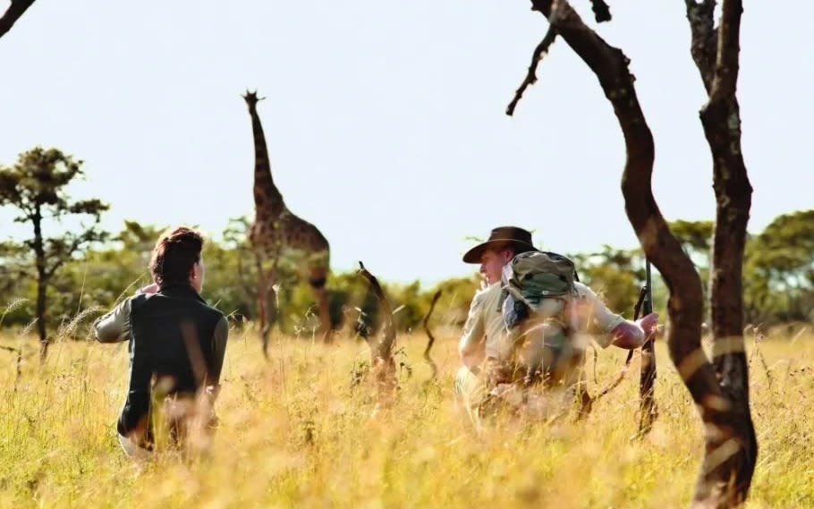 Two people squatting on an African safari with a giraffe in the background