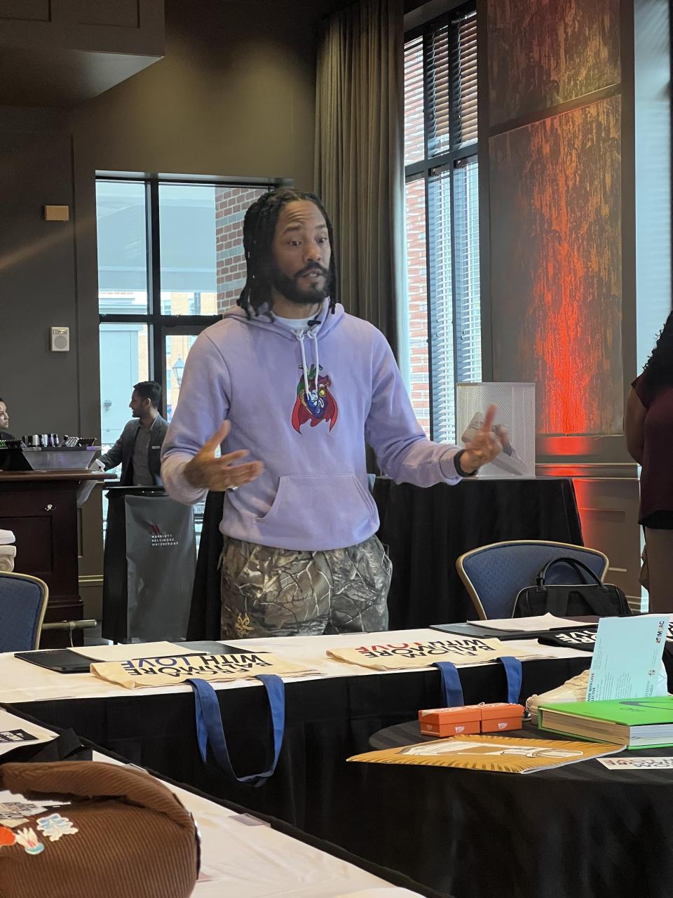 Man standing in front of a table at an indoor event, gesturing with hands, wearing a purple sweater with a graphic design