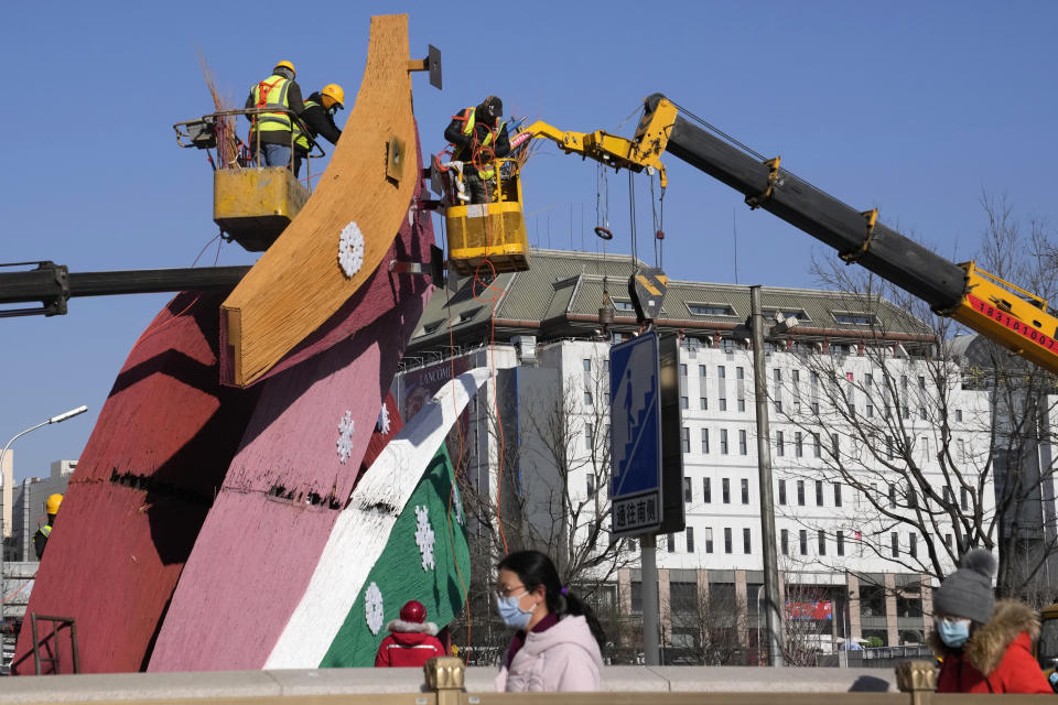 Residents wearing masks pass by workers setting up decorations with the Winter Olympics logo along a retail street in Beijing, China, Tuesday, Jan. 11, 2022. The Chinese capital is on high alert ahead of the Winter Olympics as China locks down a third city elsewhere for COVID-19 outbreak. (AP Photo/Ng Han Guan)