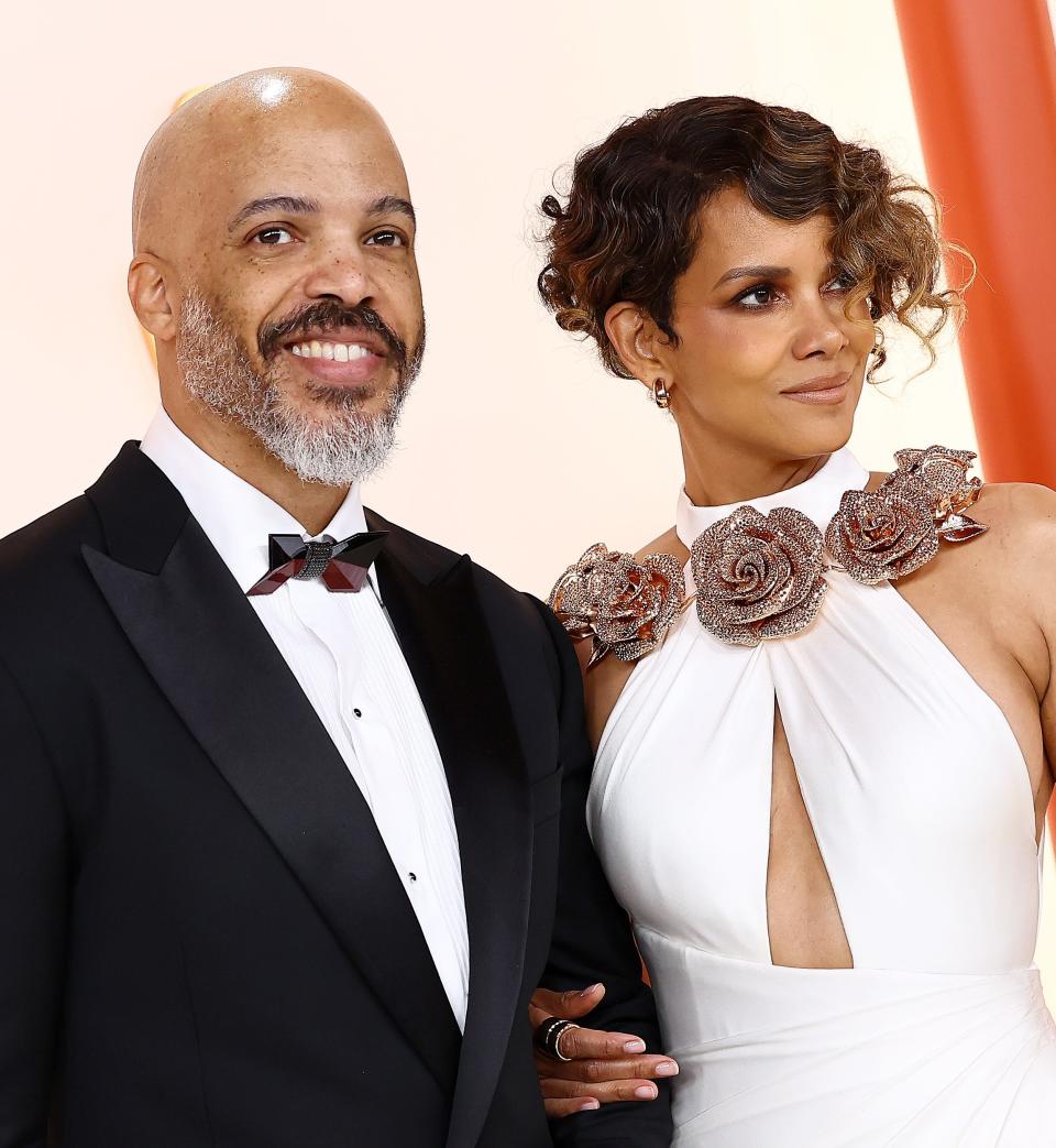 Two individuals posing at an event, with one wearing a black suit and bow tie and the other in a white dress with floral shoulder detail