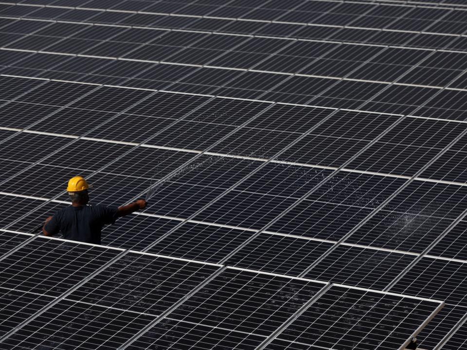 worker in yellow hard hat standing amid field of solar panels spraying water