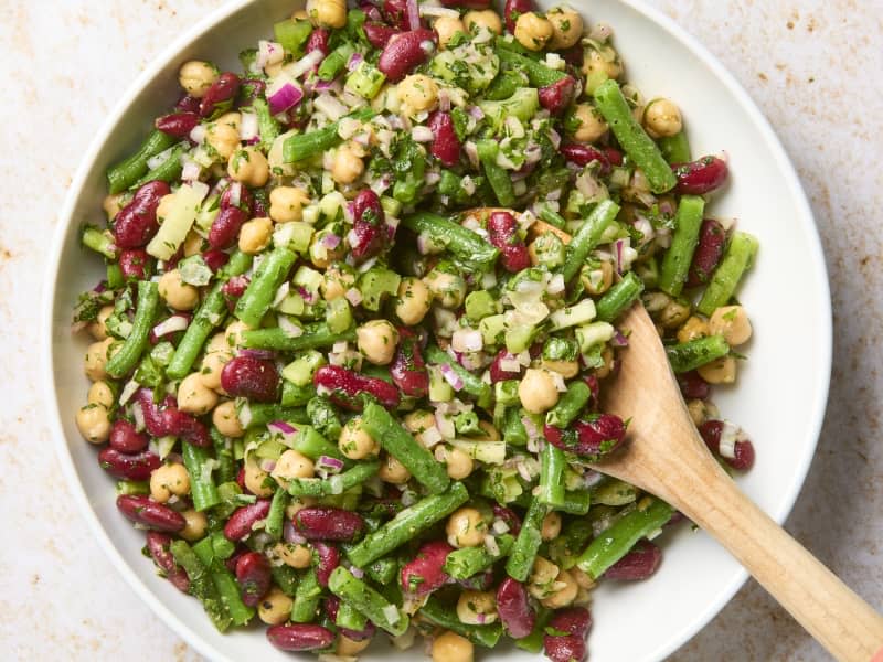 overhead shot of a 3 bean salad in a white bowl, with a wooden spoon in the bowl