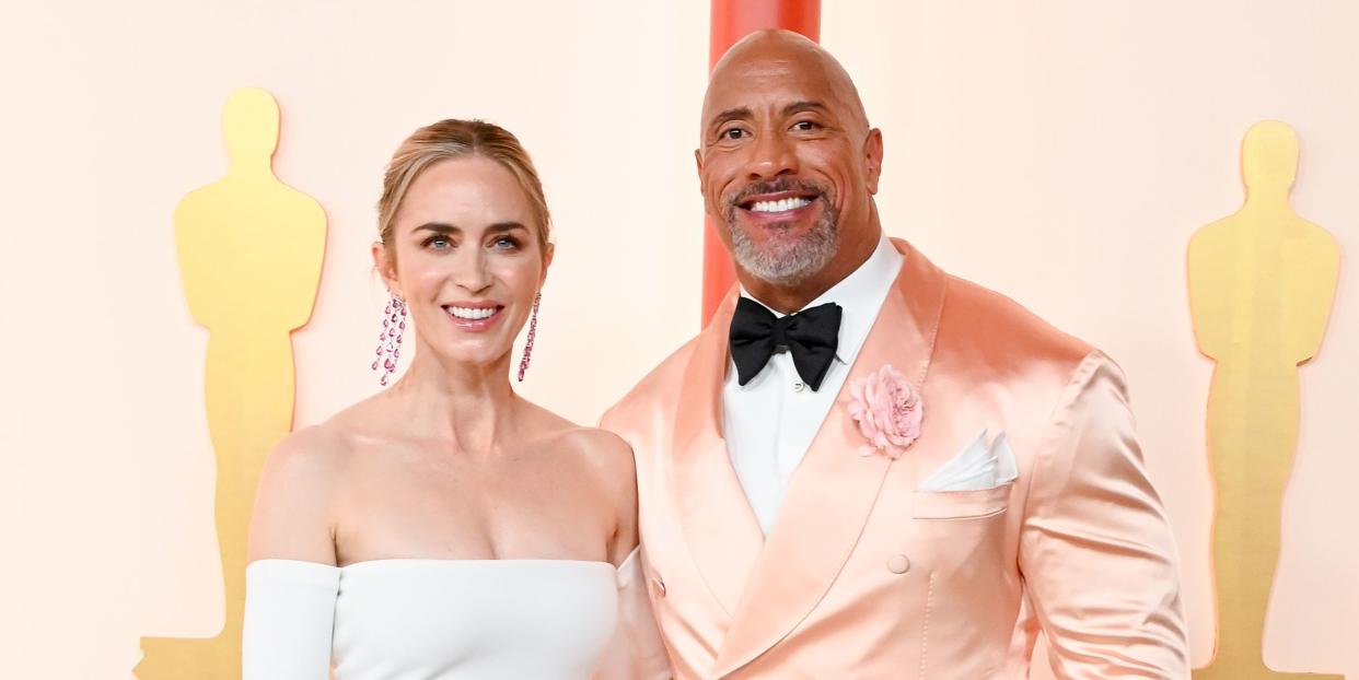 emily blunt in a strapless long white dress links arms with dwayne johnson in a black bowtie and trousers with peach satin tuxedo jacket