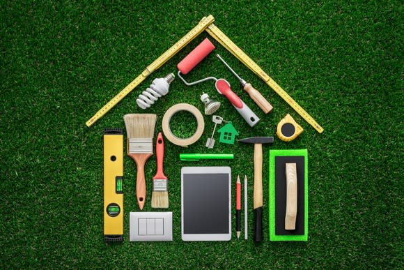 A variety of handheld home improvement tools, like paint brushes, rulers, a light bulb, and hammers, laid out neatly in the shape of a house on top of green astro turf.