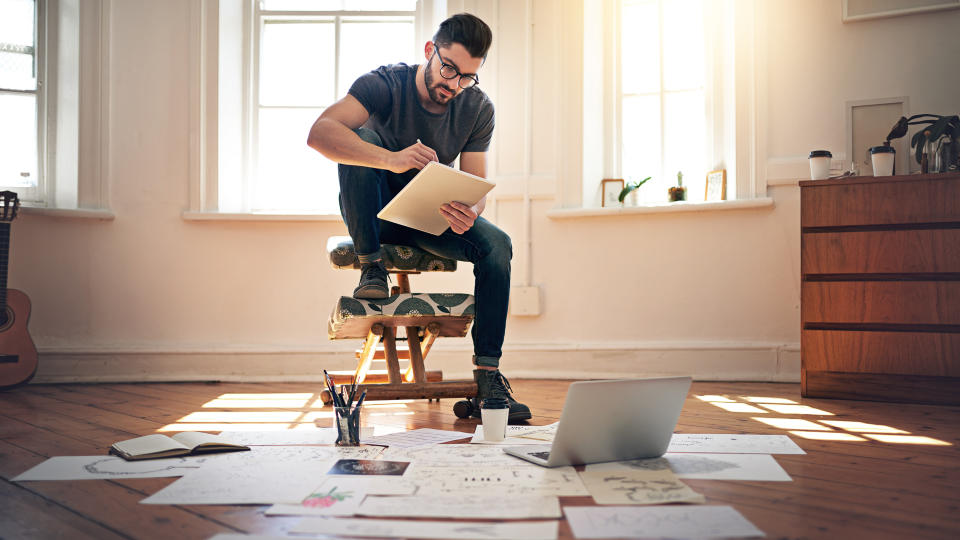 Shot of a designer working on his tablet with designs spread out on the floor in front of him.