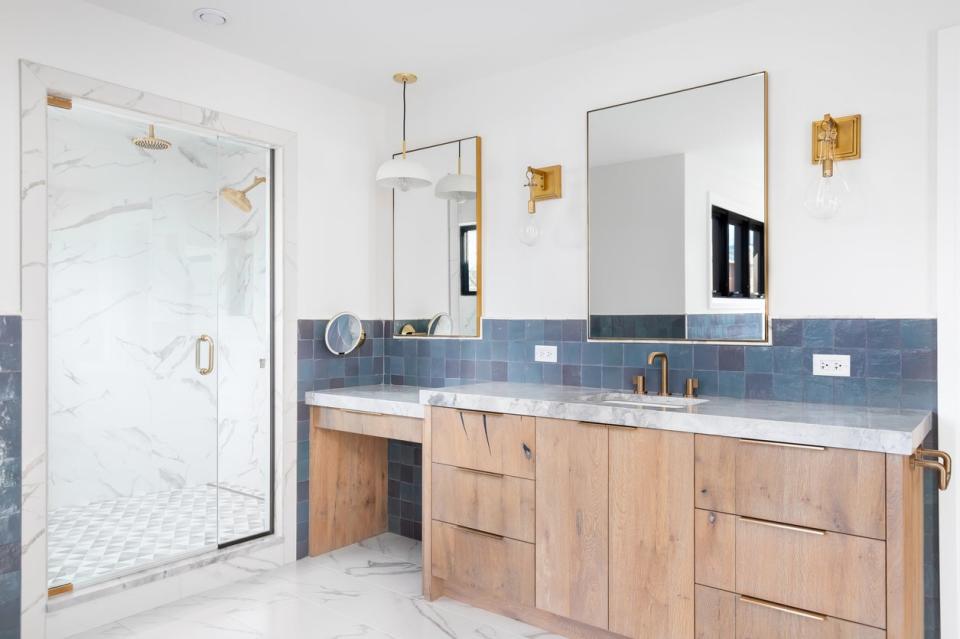 A large bathroom with blue tile that goes halfway up the wall, a wood double vanity, brass fixtures, and a luxe marble shower.