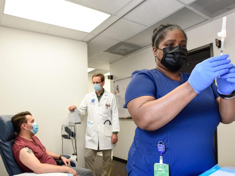 woman in scrubs prepares covid-19 vaccine, with patient and doctor chatting in background