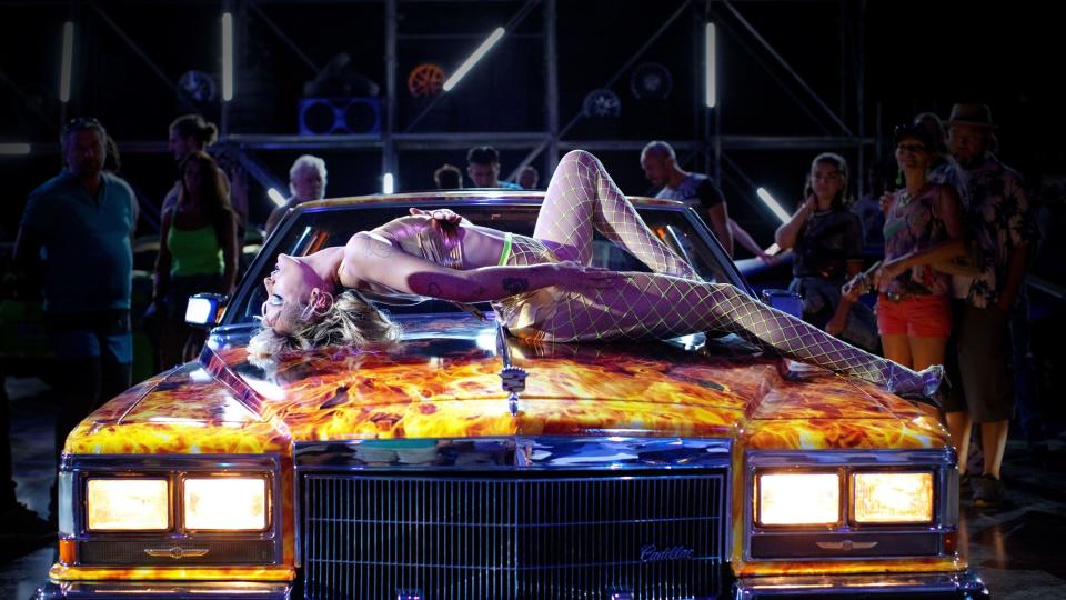 An exotic dancer rubs her leg on the hood of a flamboyant car