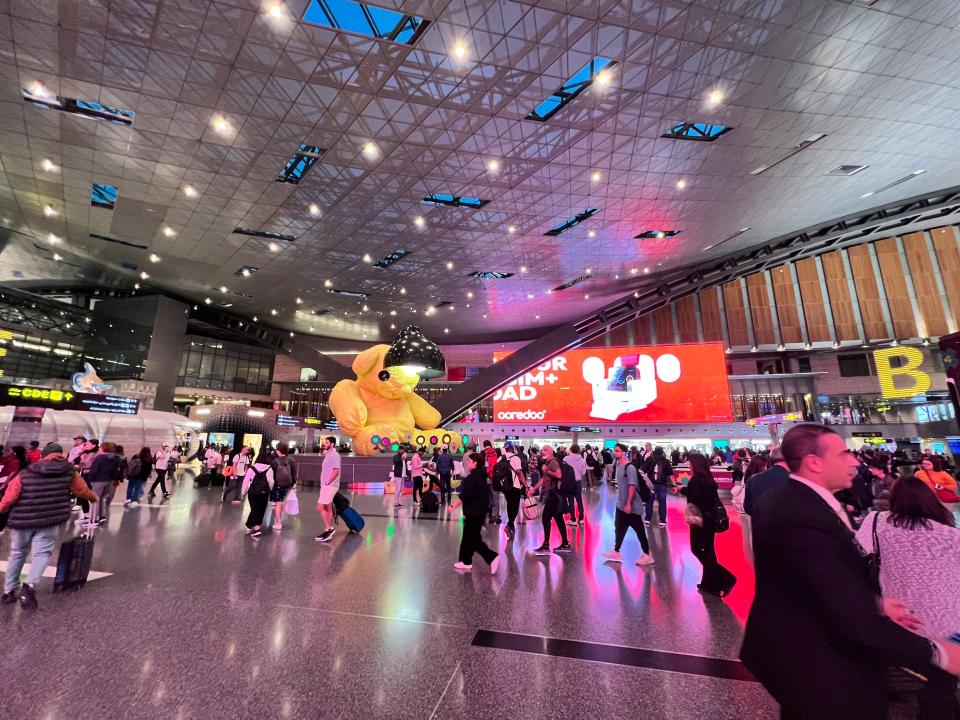 Travalers walk past a large advert screen and a statue of a yellow bar at Hamad International Airport in Doha, Qatar