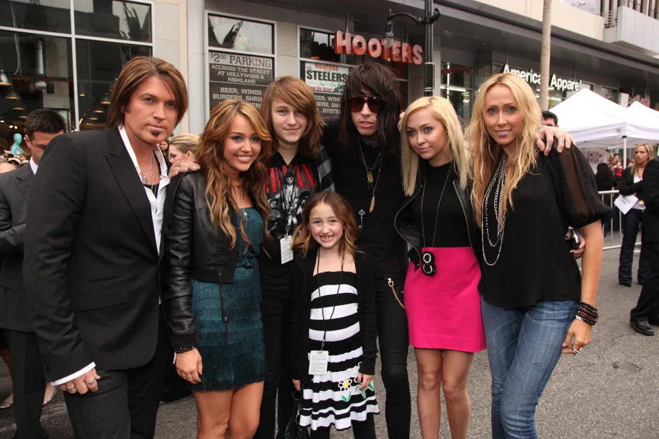 Billy Ray Cyrus, Miley Cyrus, and family pose together, with some dressed in casual attire and others in black and pink accents, at an outdoor event