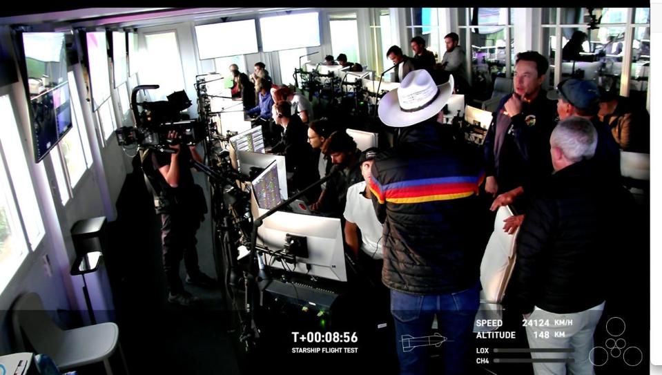 elon musk talks to man in cowboy hat in spacex control room full of computers and flight controllers