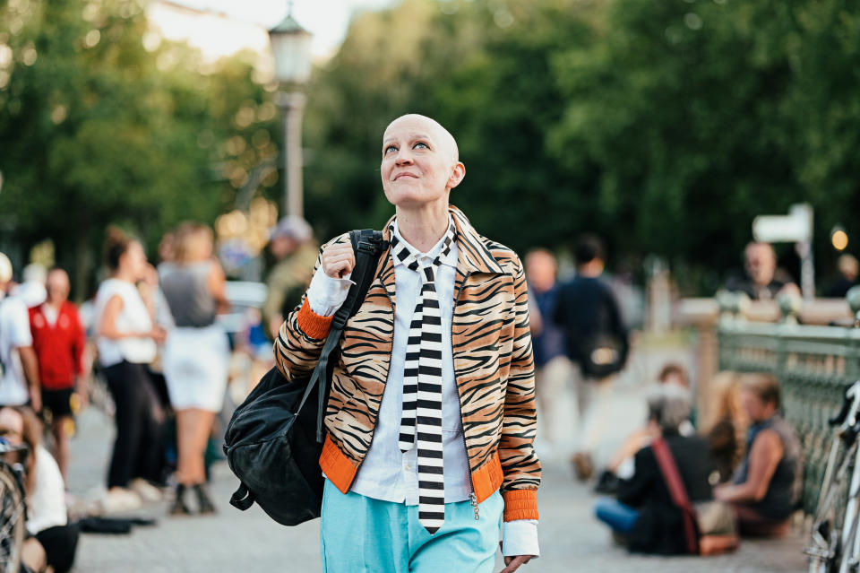 Person with a bald head wearing a patterned jacket, striped tie, and blue pants, carrying a backpack, looks up while walking in a busy outdoor area