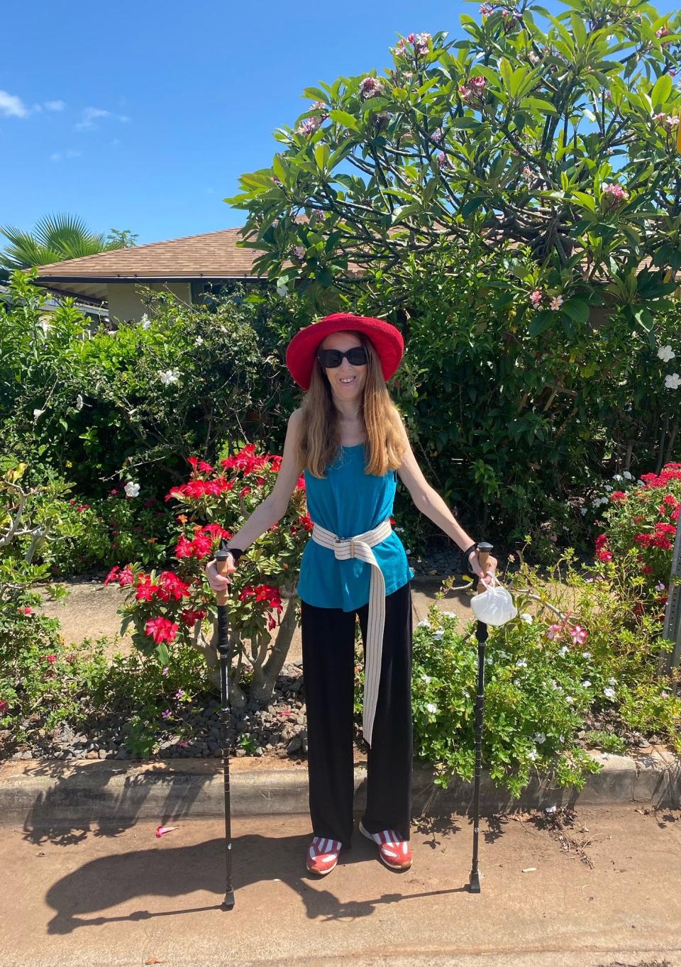 Antonia Hall poses wearing a blue T-shirt and black pants near her home in Maui, Hawaii, with hiking sticks to support her while she walks.