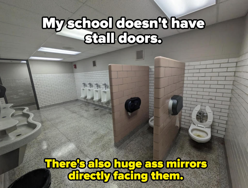 An empty men's restroom with urinals on the far wall, two stalls, and a multi-sink area. The stall toilets have open lids, and there are light fixtures on the ceiling