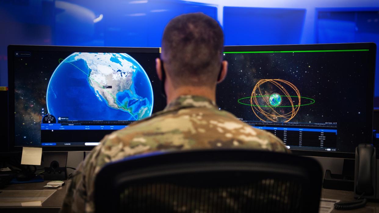  A person in camouflage sits at a computer screen looking at an animation of several rings around Earth denoting spacecraft orbits . 