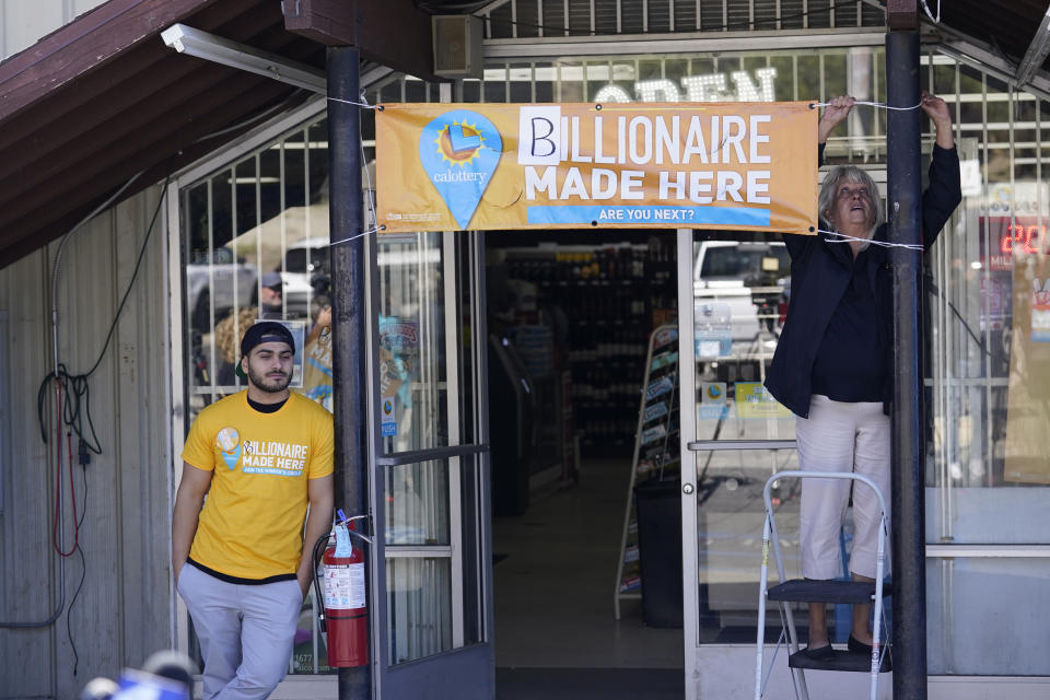 Lottery official Anita Ayers, right, hangs a banner above the entrance to the Midway Market & Liquor store where a winning lottery ticket was sold in Frazier Park, Calif., Thursday, Oct. 12, 2023. The liquor store in a tiny California mountain town reverberated with excitement after word that the winning ticket for a $1.765 billion Powerball jackpot was sold there. The drawing Wednesday night ended a long stretch without a winner of the top prize. (AP Photo/Marcio Jose Sanchez)