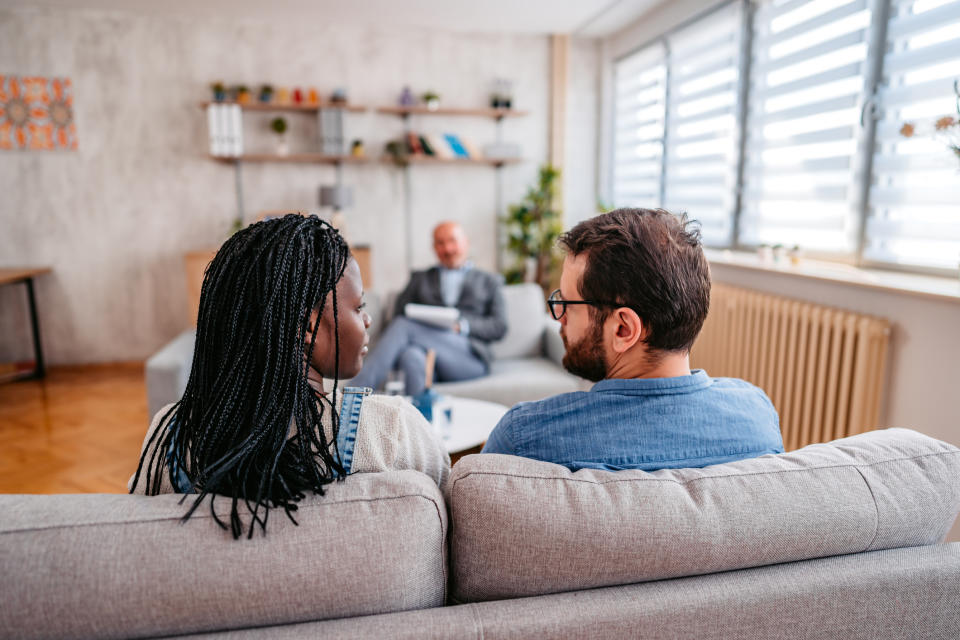 A couple sits on a couch facing a therapist in an office setting. Shelves with books and decor are in the background