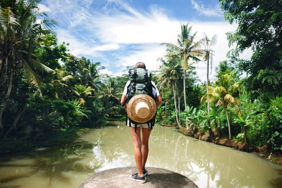 Woman with backpack standing on the edge of a big tropical river and looking far away