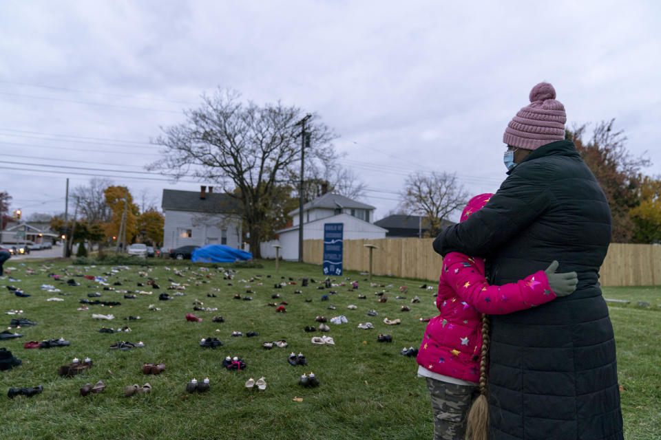 Theresa McGarity, left, embraces her granddaughter, Blake, 8, while remembering her mother, Carolyn Wilson, during an art installation vigil with shoes representing residents in Macomb County who died from COVID-19 in Mount Clemens, Mich., Friday, Oct. 30, 2020. Wilson, who died in April at 76 years old, was a God-fearing woman and had been teaching Blake how to crochet. On Friday night, as McGarity brought Blake to the art installation, a raucous pro-Trump demonstration down the street could be heard as they read the age and hometown of each of their dead. (AP Photo/David Goldman)