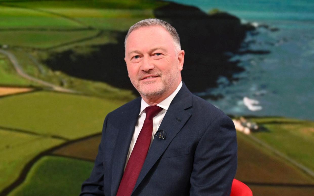 Steve Reed smiles in front of a backdrop showing a stretch of coastline