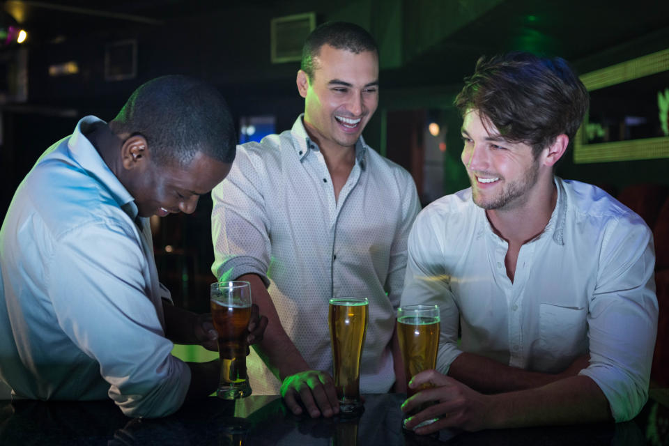 Three men at a bar laughing and talking, each holding a glass of beer. The setting appears to be a lively social environment. Names of persons not available