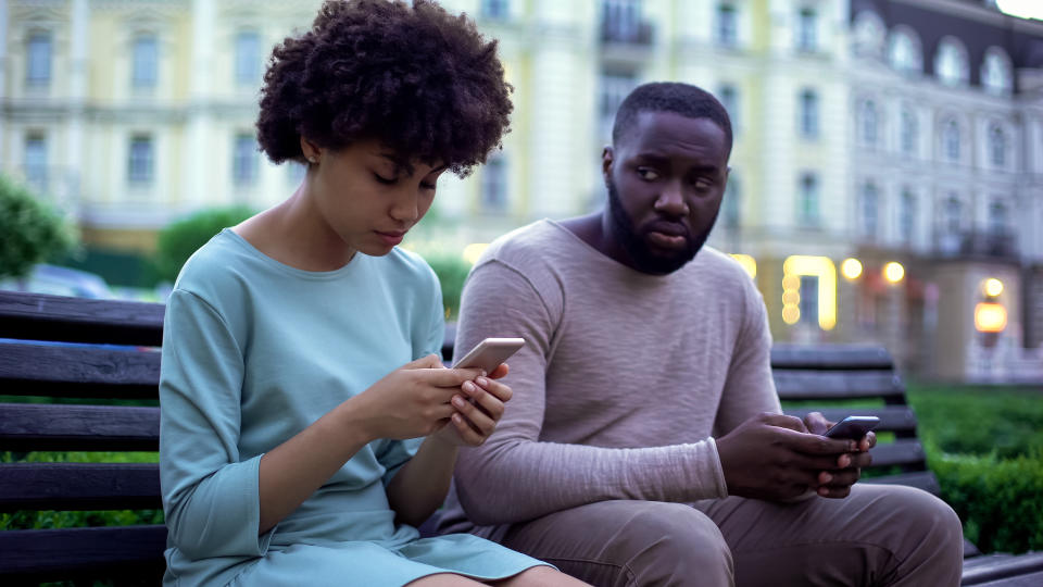 A woman in a casual dress and a man in a casual sweater, sitting on a bench, both looking at their phones with different expressions