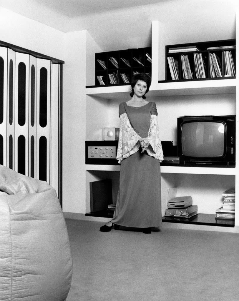 A woman stands in a modern living room with shelving behind her stocked with records and a television. She wears a long dress with lace sleeves