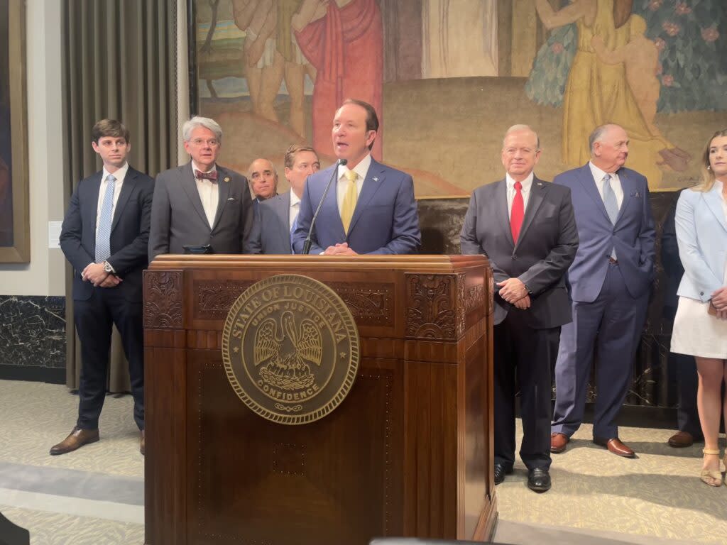 A man in a suit speaks at a podium while other people stand behind him