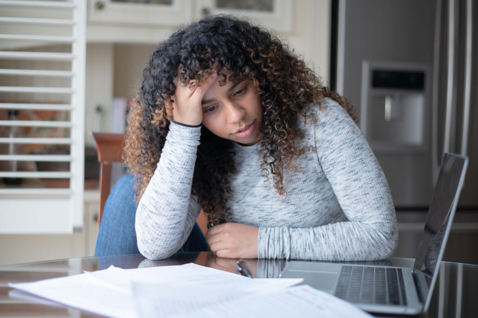 A mixed race woman sit at a table with one knee bent up on her chair.  She has her head resting in her hand with her elbow on the table and is looking frustrated.  Shed is wearing casual clothing.