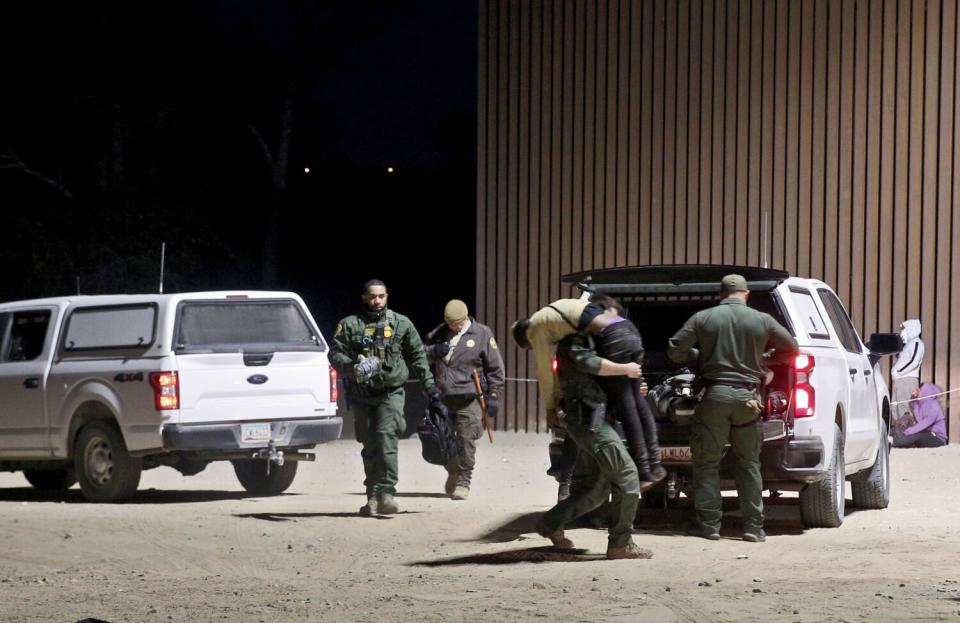 A Border Patrol officer carries a man for help as other officers mill around two white pickup trucks parked in the dirt