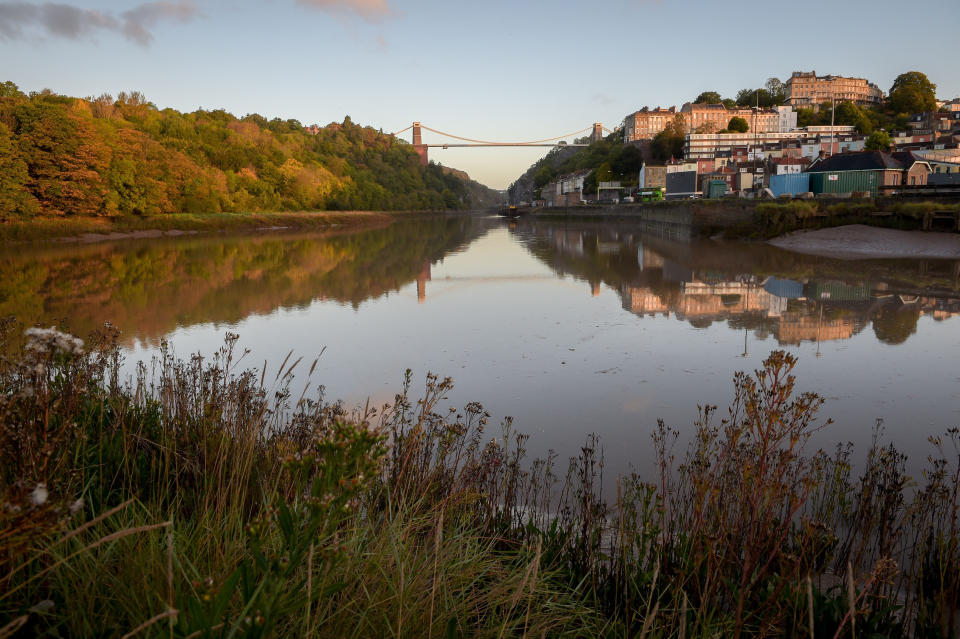 Sun rises over the Avon Gorge as the Clifton Suspension Bridge and Autumnal trees are reflected in the River Avon, Bristol.