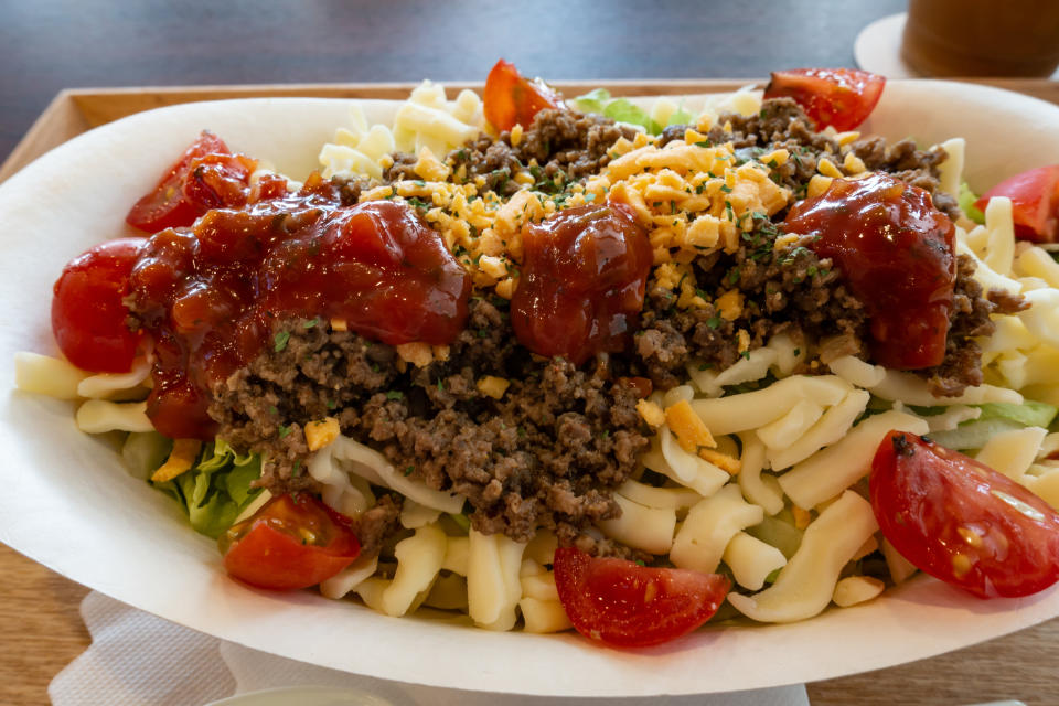 A dish featuring ground beef, shredded cheese, tomatoes, and a drizzle of red sauce, garnished with chopped nuts, served in a paper container