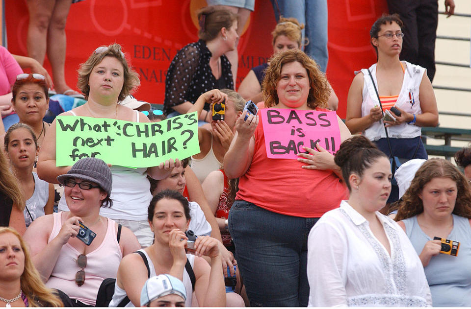 People in an audience with some holding signs, including "Bass is back!" and "What's next for Chris's hair?"