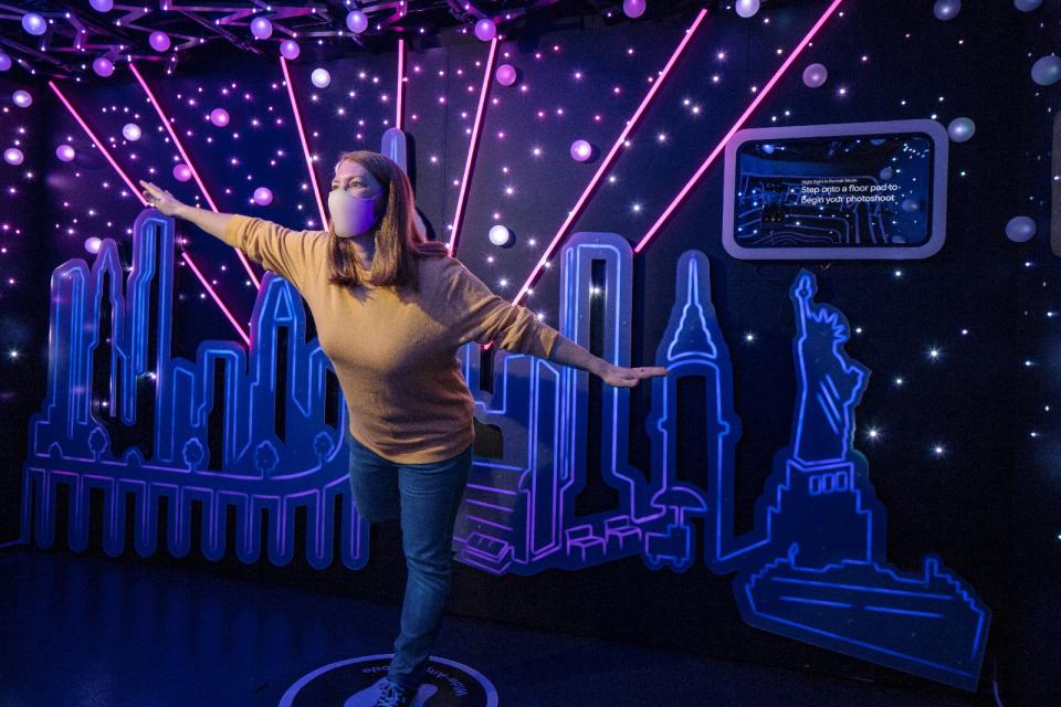 <p>Google Store Chelsea. A Night Sight-themed Sandbox with a person wearing a face mask posing in the middle. The background features a cut-out art rendering of the New York City skyline with rays of pink light emanating from it and the Statue of Liberty on the right. A display is on the wall above this. </p>
