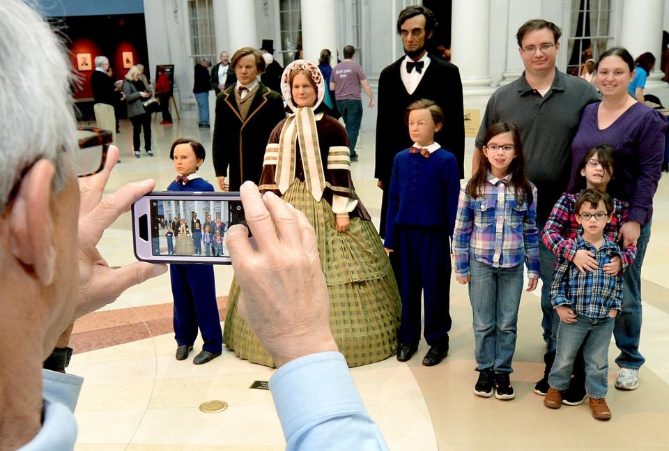Abraham Lincoln Presidential Library and Museum volunteer Larry Stone takes a photo of a family with their phone camera as they stand  in front of the Lincoln family figurers in the plaza of the Lincoln museum Saturday Feb. 11, 2023.