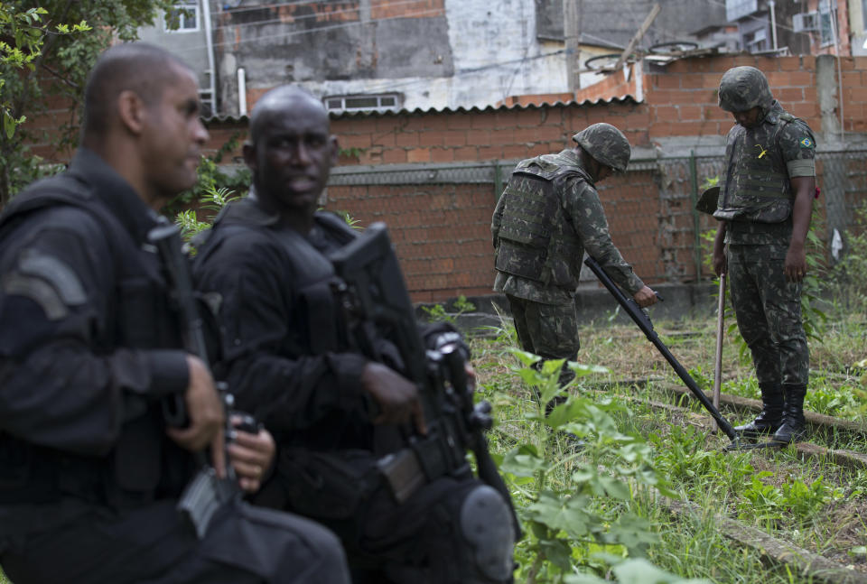 Special Police Operations Battalion (BOPE) officers stand guard, as army soldiers look for weapons with the aid of a metal detector during an operation in the Mare slum complex, ahead of its "pacification," in Rio de Janeiro, Brazil, Wednesday, March 26, 2014. Elite federal police and army troops will be sent to the city to help quell a wave of violence in so-called "pacified" slums. Recent attacks on police bases in the favelas is raising concerns about an ambitious security program that began in 2008, in part to secure the city ahead of this year's World Cup and the 2016 Olympics. (AP Photo/Silvia Izquierdo)