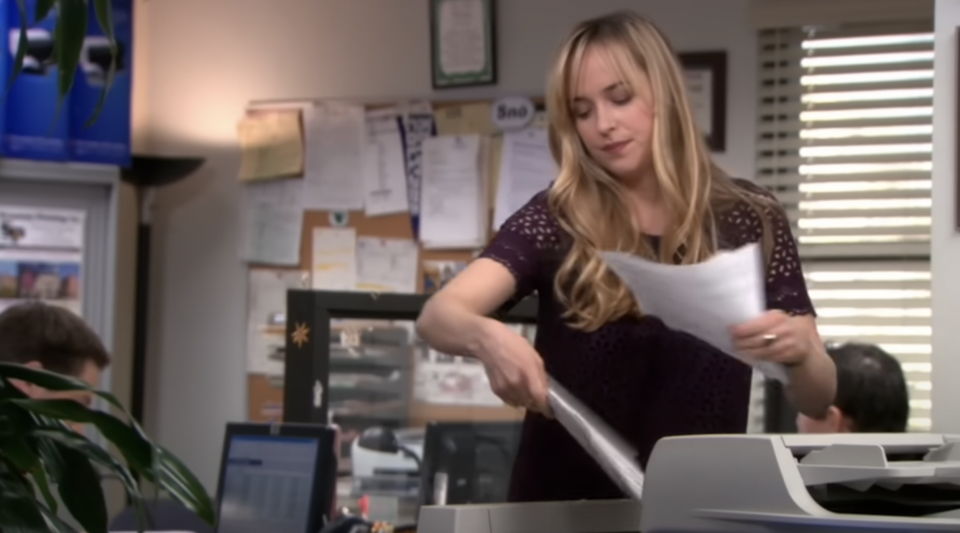 Woman in office attire sorting papers by a printer, with a colleague seated nearby. Scene from TV show